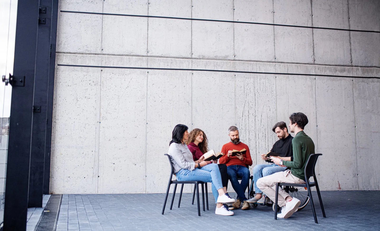 Five adults sit in a circle of chairs against a concrete wall, reading and discussing books together, one of them in a wheelchair