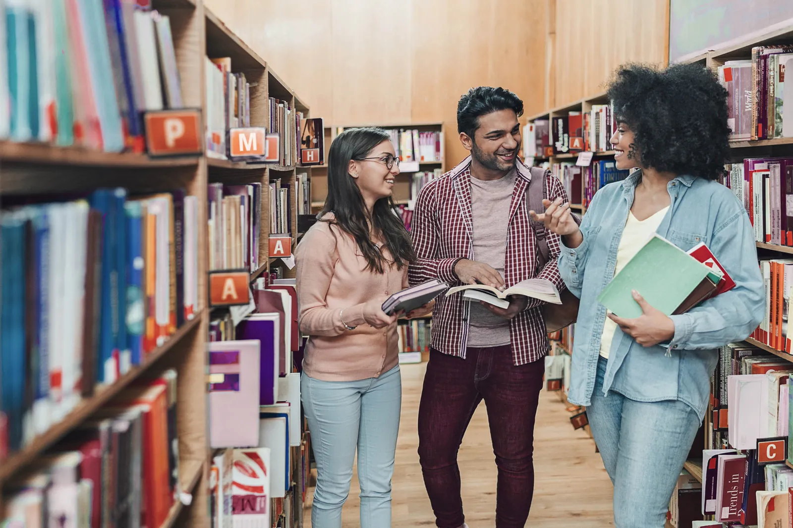 Three smiling friends chat between tall library shelves, each holding an open book or a stack of books