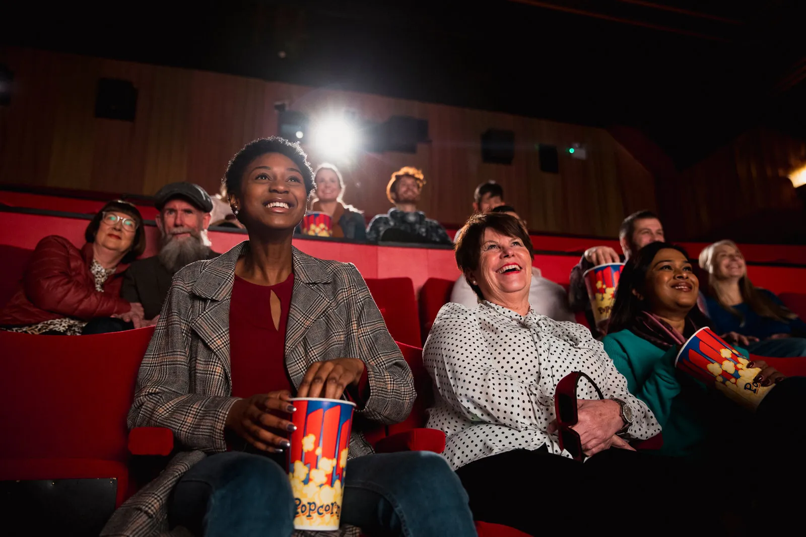 Smiling audience in red cinema seats watching a film, several holding striped popcorn tubs, with the projector light behind them