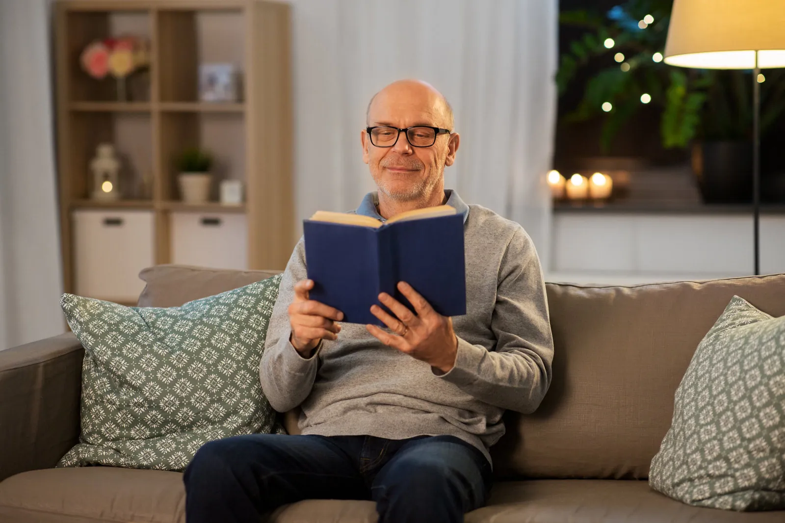 An older man in glasses sits on a sofa at home in the evening, smiling as he reads a blue hardback book