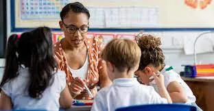 A teacher in glasses sits at a classroom table helping young pupils with their work