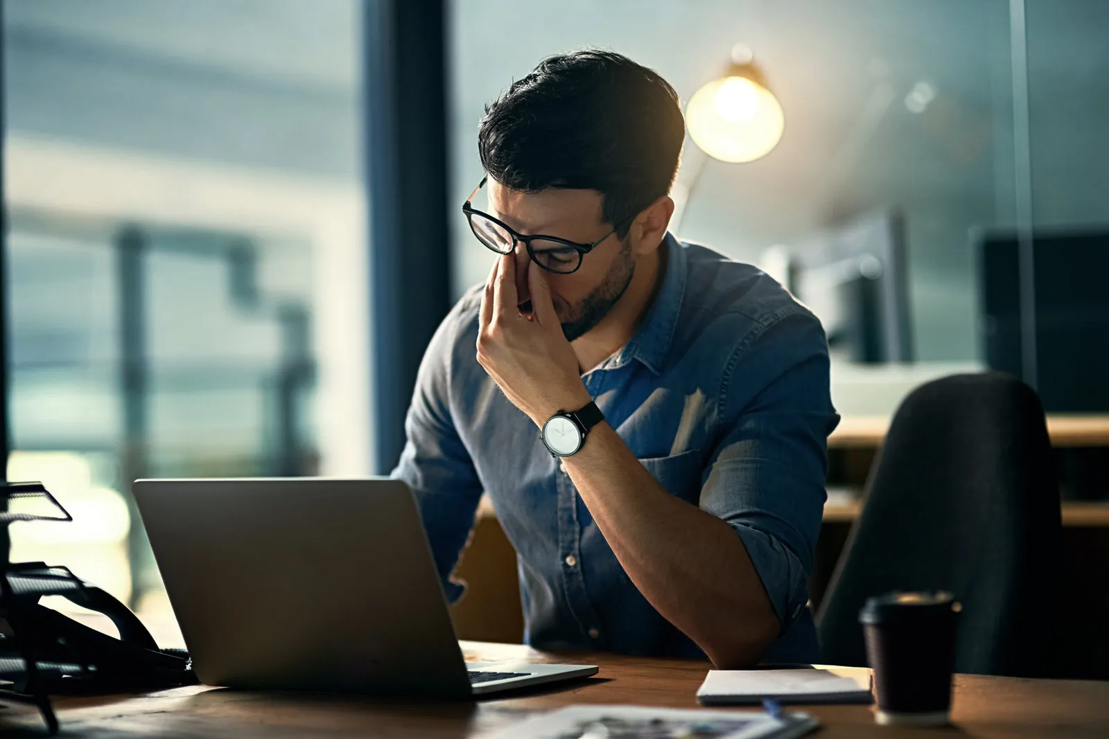 A tired man in glasses rubs his eyes at a laptop in a dim office at night, a coffee cup beside him