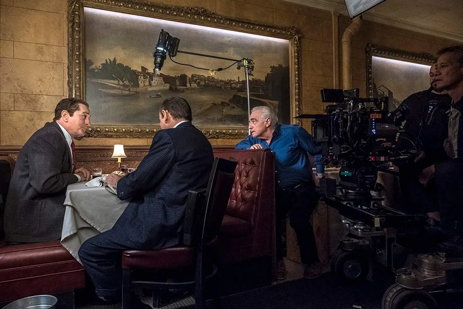 A white-haired director in a blue shirt leans on a red booth, watching two suited actors talk at a restaurant table beside a camera rig