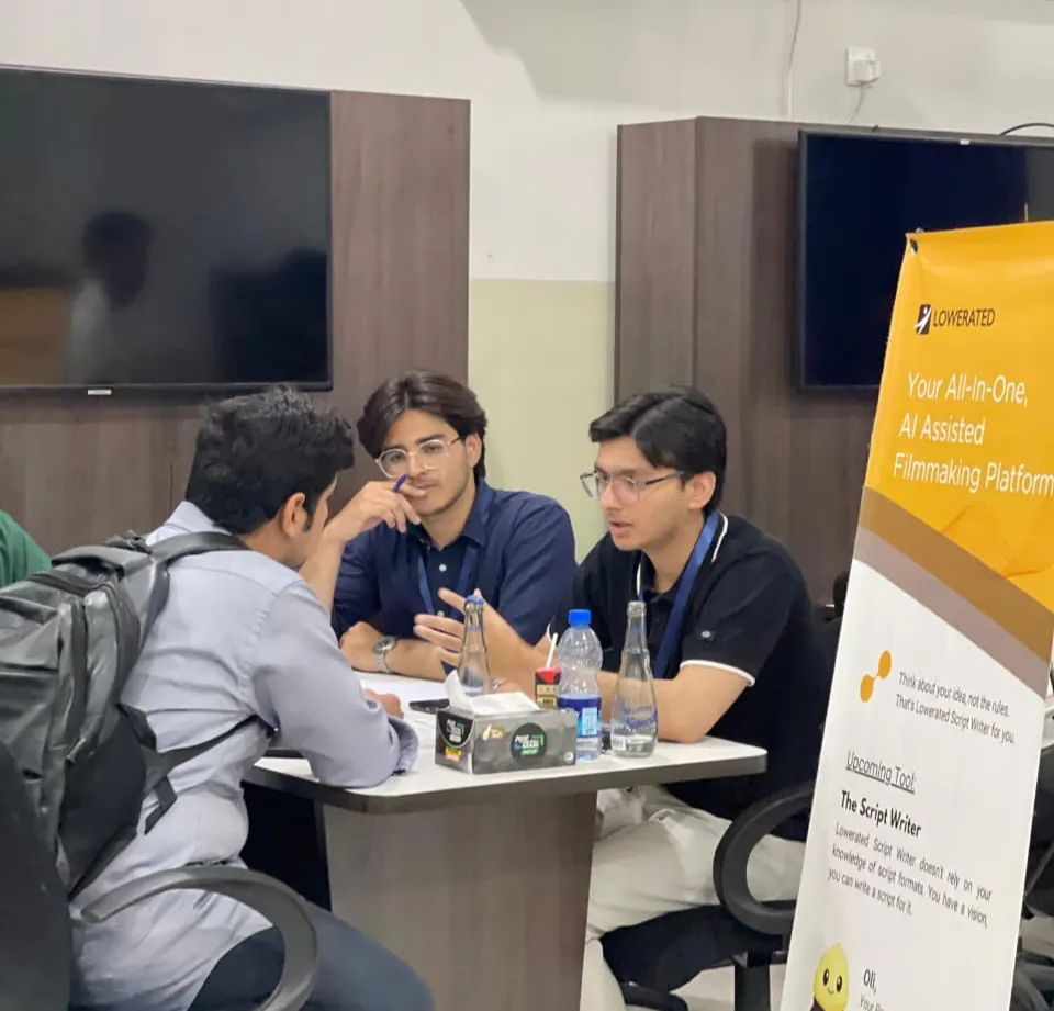 Two young Lowerated team members in glasses talk with a visitor at an expo table, beside a yellow 'AI Assisted Filmmaking Platform' banner