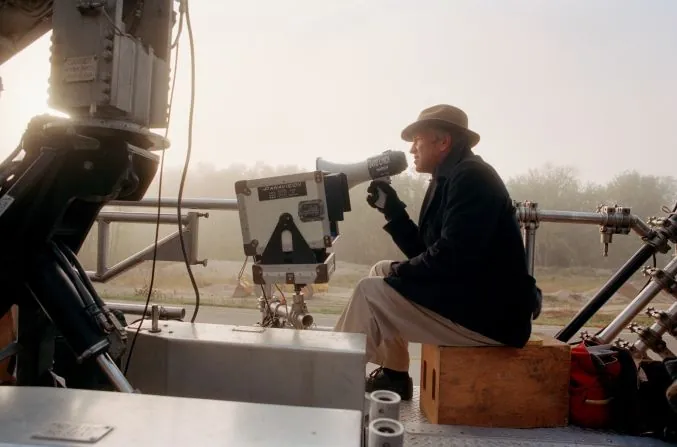 A director in a hat and dark coat sits on an apple box beside a camera rig, speaking through a megaphone on a misty outdoor set