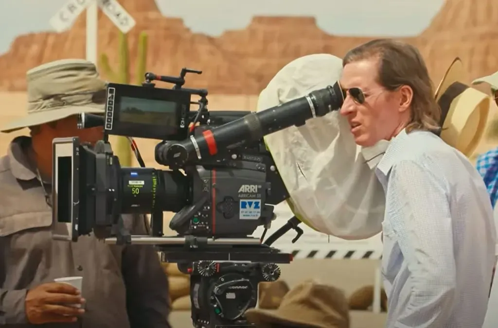 A director in sunglasses looks through an ARRI film camera on a desert film set with a painted backdrop of mesas behind